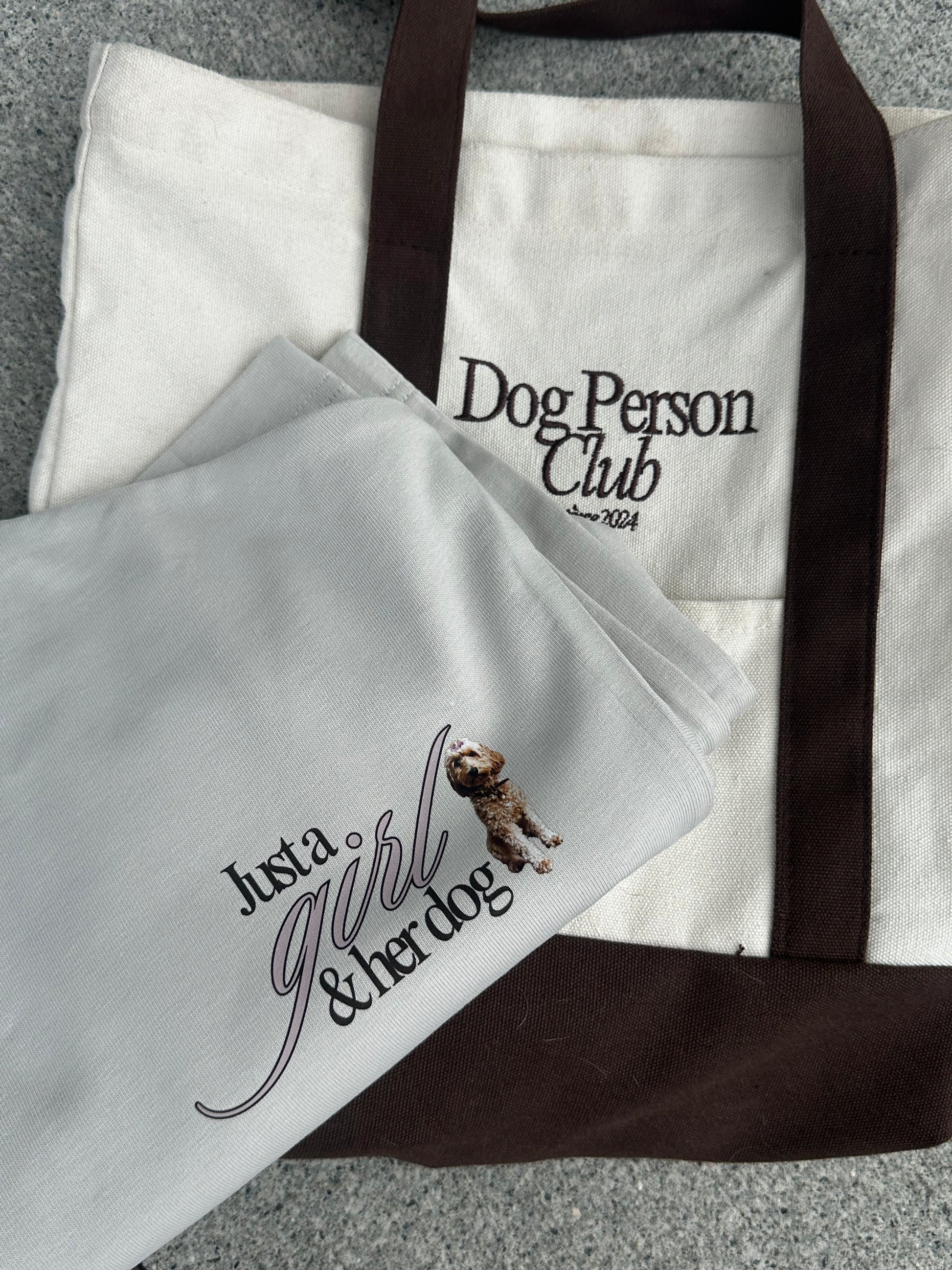 Close-up of a cream “Just a girl & her dog” tee laid over a cream and brown Dog Person Club tote bag.
